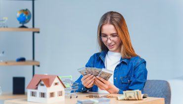 Young pretty girl counts her savings money and looks to a model of the house.|The parent teaches the child how to handle money. Financial literacy course for children of preschool and school age.